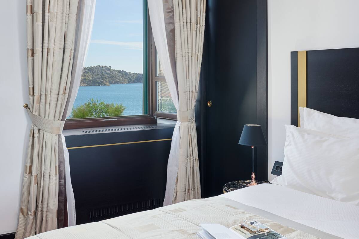 A hotel room with a bed, a window with curtains, a nightstand with a lamp, and a view of a body of water.