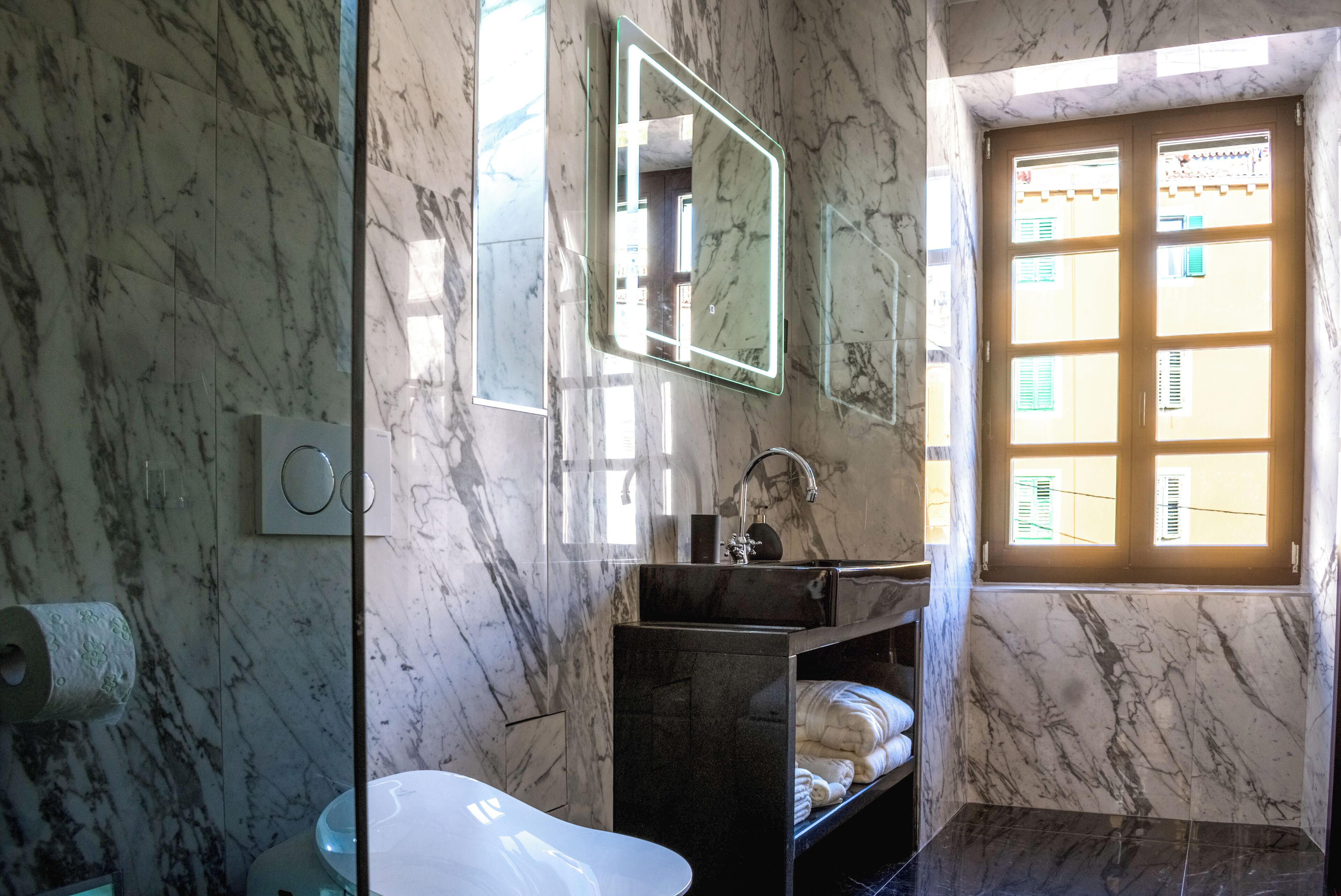 Une salle de bain avec un mur en marbre, une baignoire blanche, un miroir et une fenêtre avec des stores.