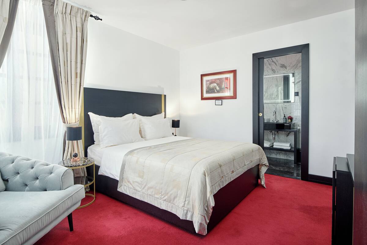 Ein Hotelzimmer mit einem Bett, einem Nachttisch mit Lampe, einem roten Teppich, einem grauen Sofa und einem durch eine Glastür einsehbaren Badezimmer.