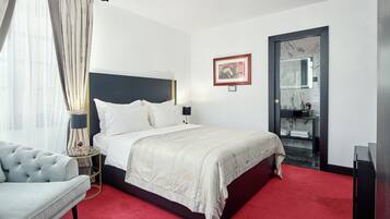 A hotel room with a bed, a bedside table with a lamp, a red carpet, a grey sofa, and a bathroom visible through a glass door.