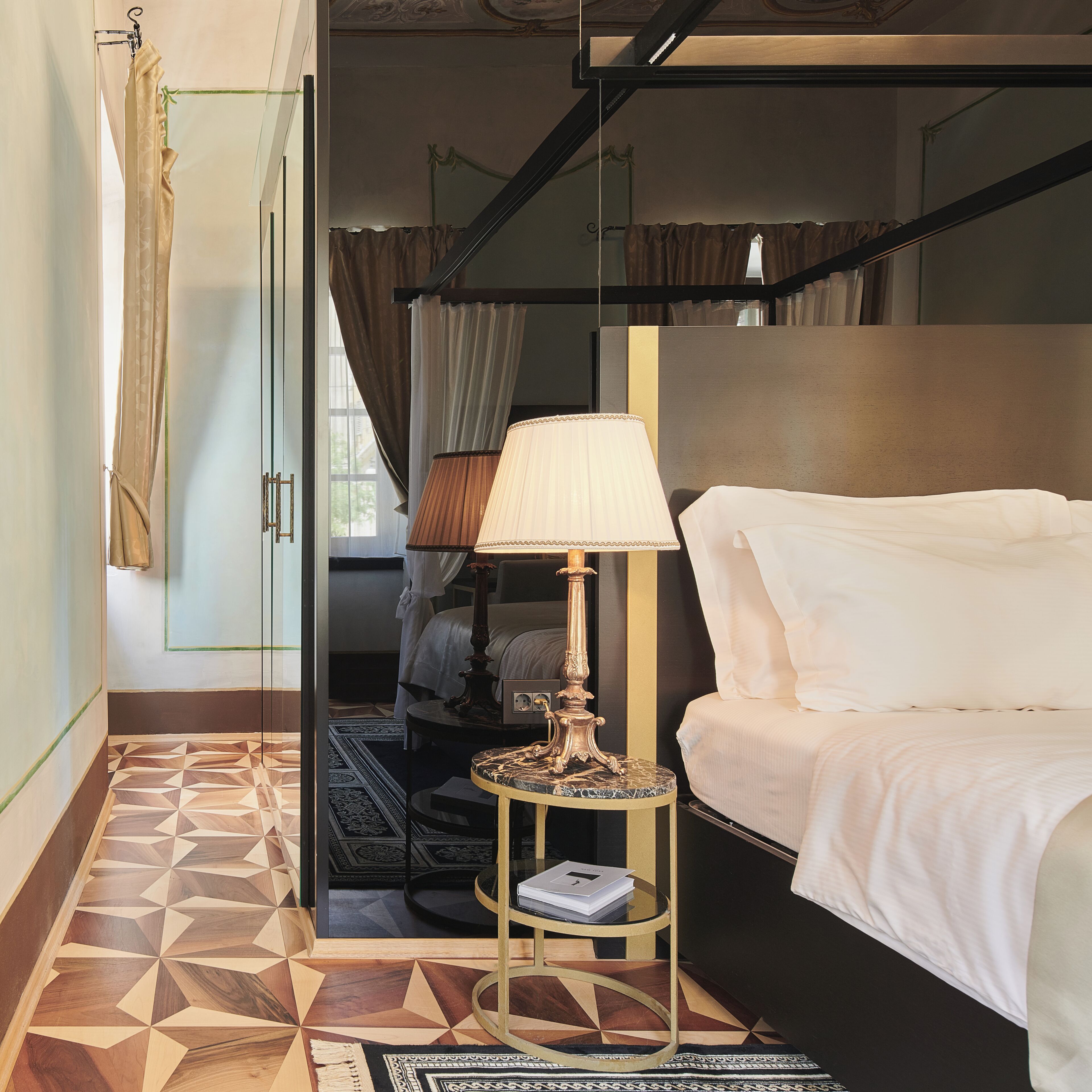 Een hotelkamer met een bed, een nachtkastje met een lamp, een spiegel en een vloer met een geometrisch patroon.