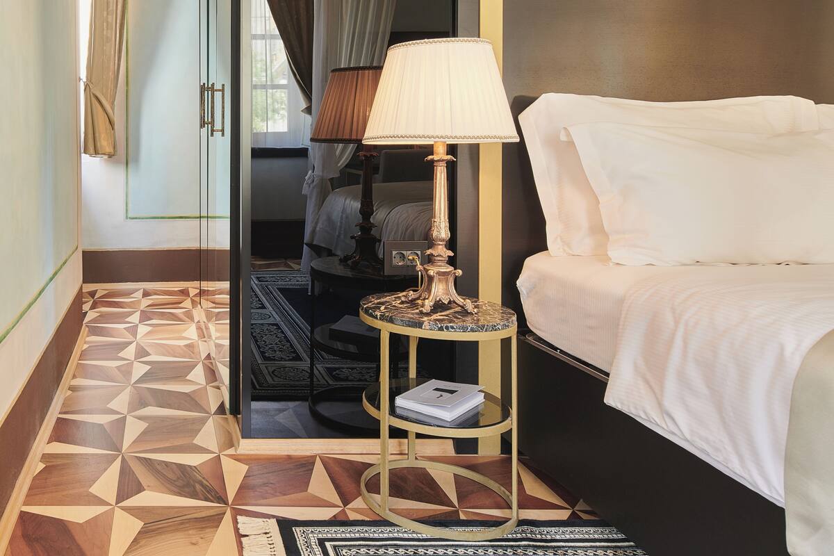 A hotel room with a bed, a bedside table with a lamp, a mirror, and a geometric patterned floor.