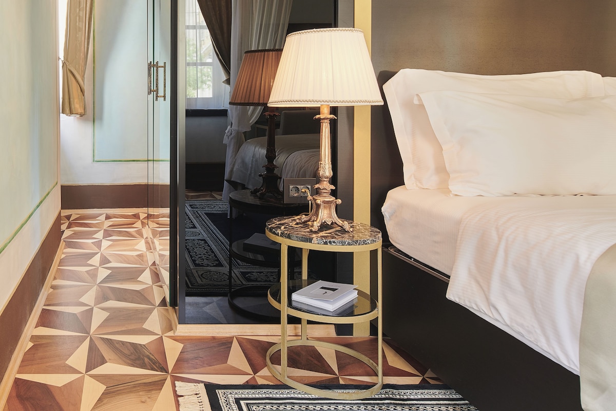 Ein Hotelzimmer mit einem Bett, einem Nachttisch mit Lampe, einem Spiegel und einem geometrisch gemusterten Boden.