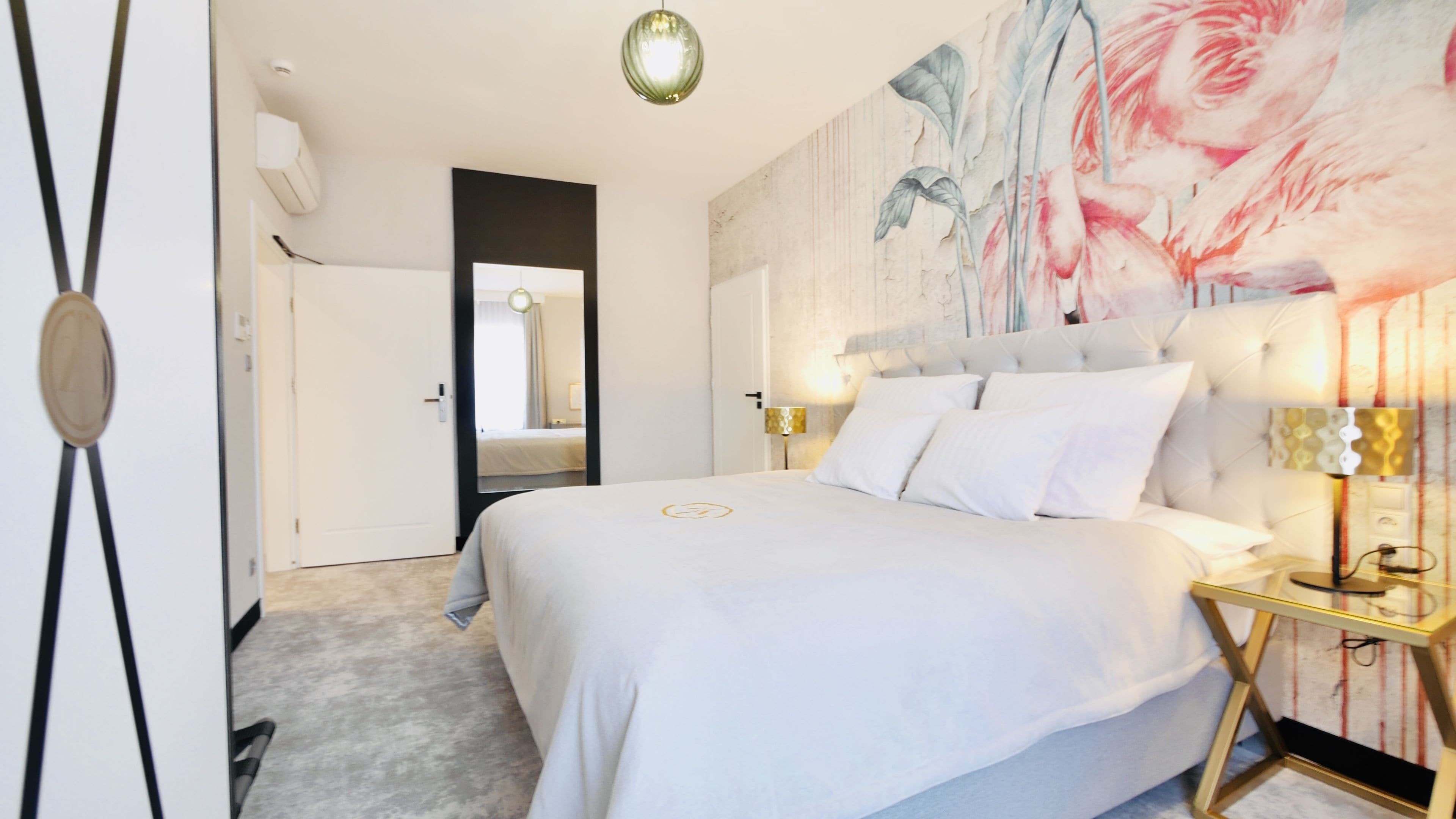A modern hotel room with a large bed, a headboard with a floral mural, bedside lamps, and a view of the hallway.