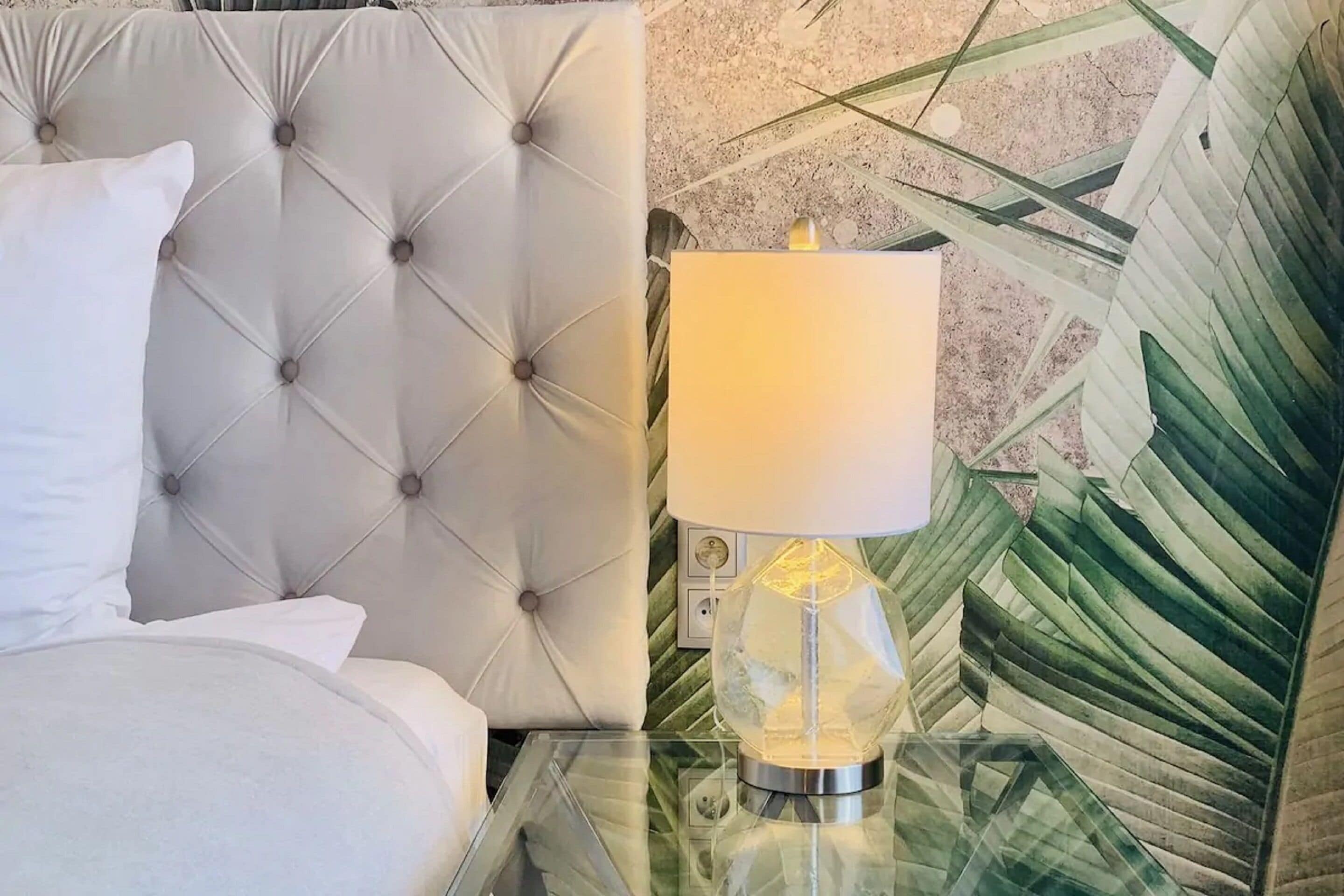 A bed with a tufted headboard, a bedside table with a glass top, and a lamp with a clear glass shade.