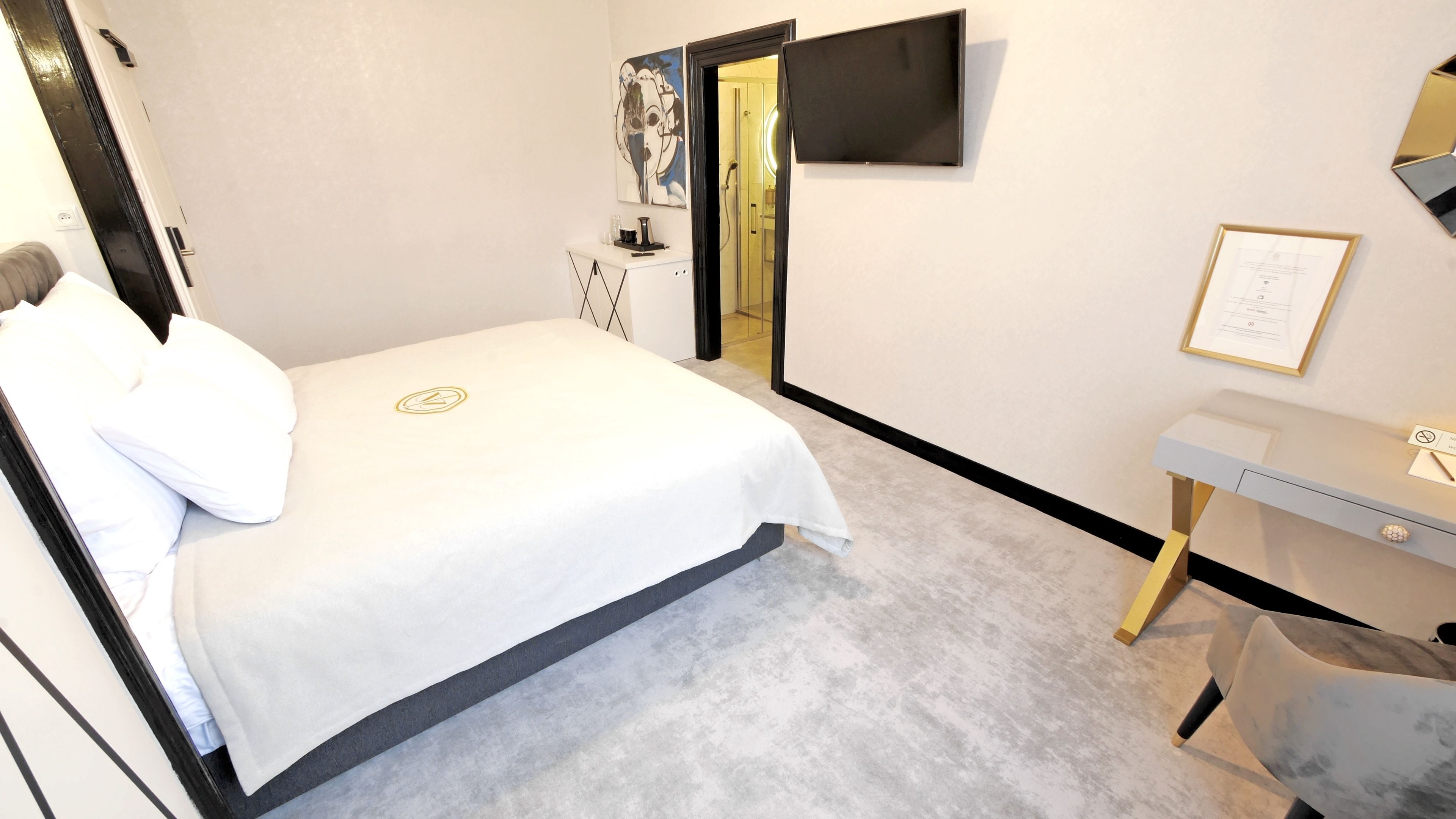 A modern hotel room with a bed, a desk, a chair, a TV, and a framed certificate on the wall.