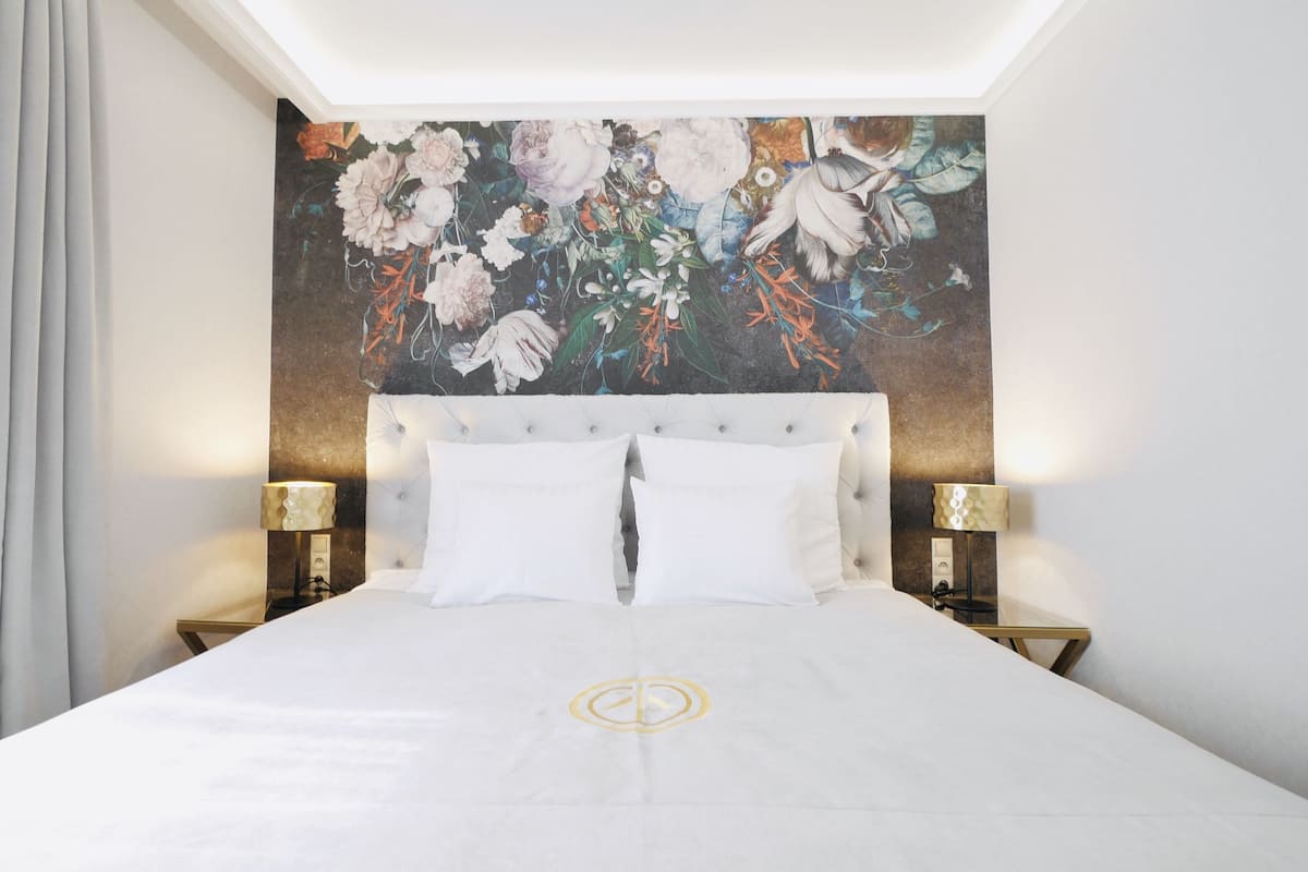 Habitación de hotel con una cama grande, dos mesitas de noche con lámparas, un mural floral en la pared y una ventana con cortinas.