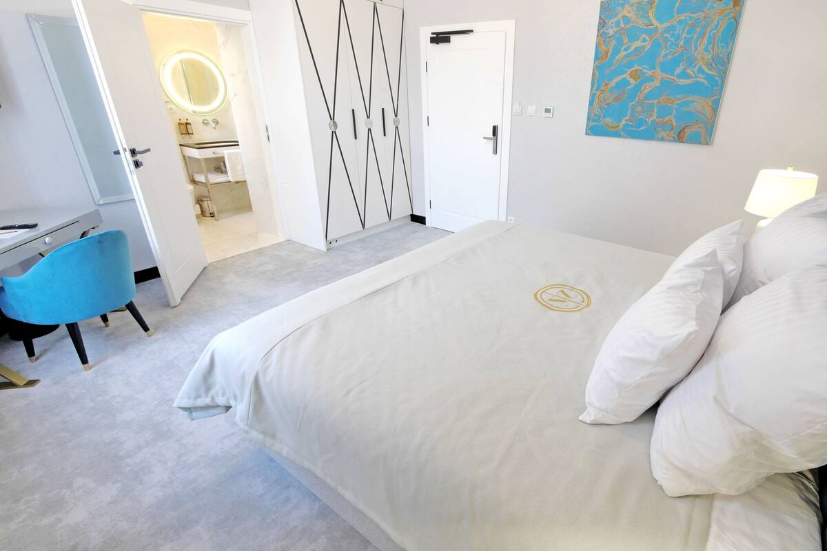 Un dormitorio con cama, escritorio, silla y baño visible a través de una puerta abierta.
