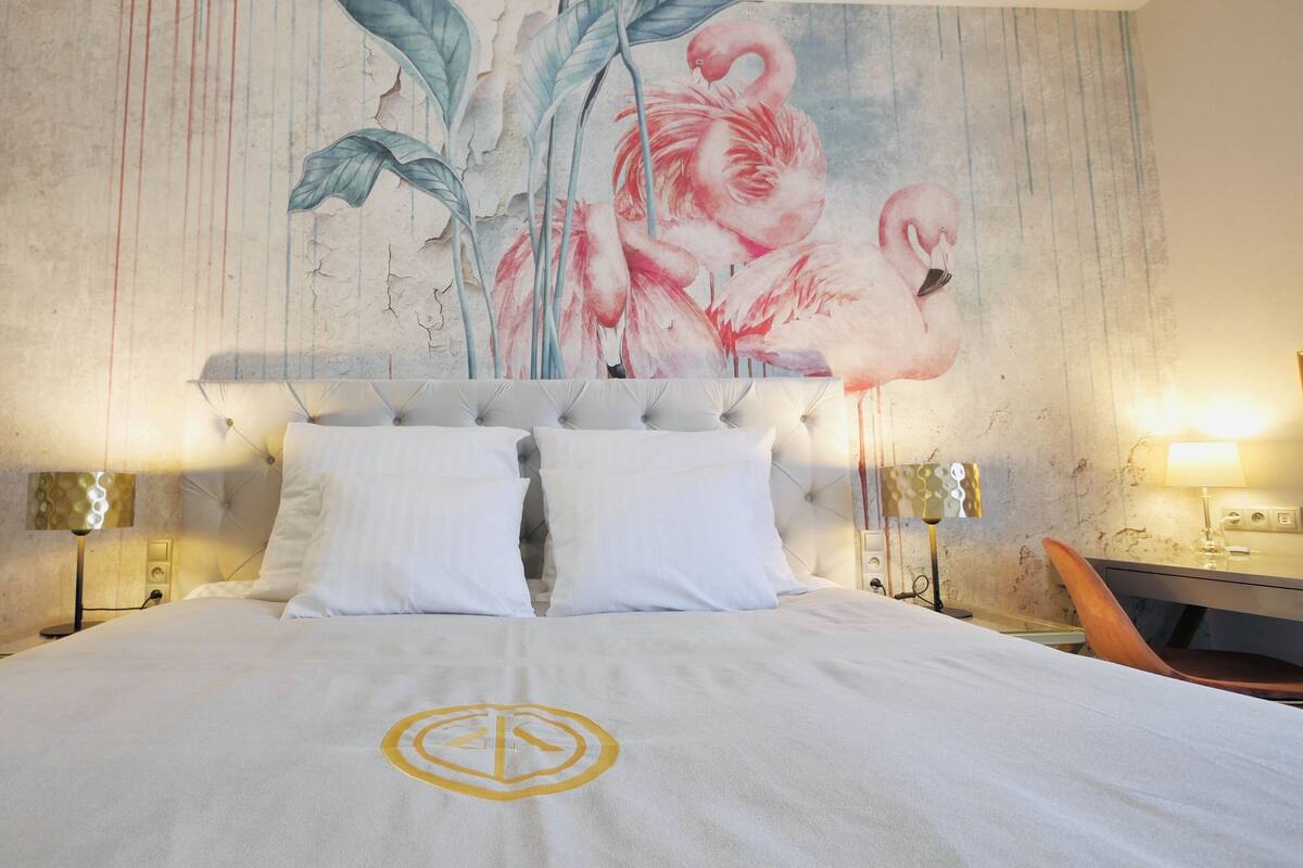 Habitación de hotel con una cama grande, ropa de cama blanca, un cabecero con un mural floral, dos lámparas de noche y una silla de madera.