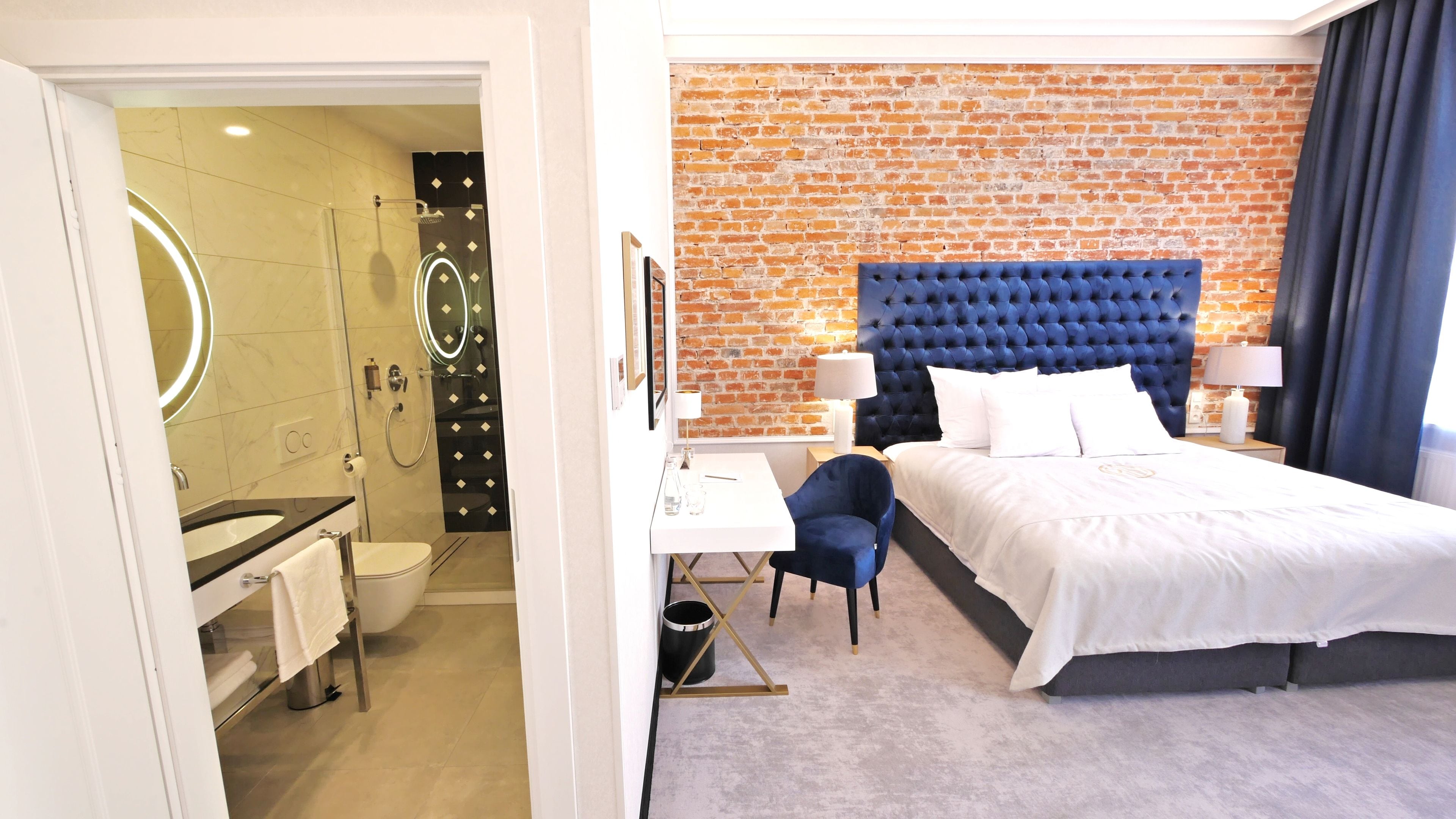 A hotel room with a modern bathroom, a bed with a tufted headboard, and a desk with a chair.