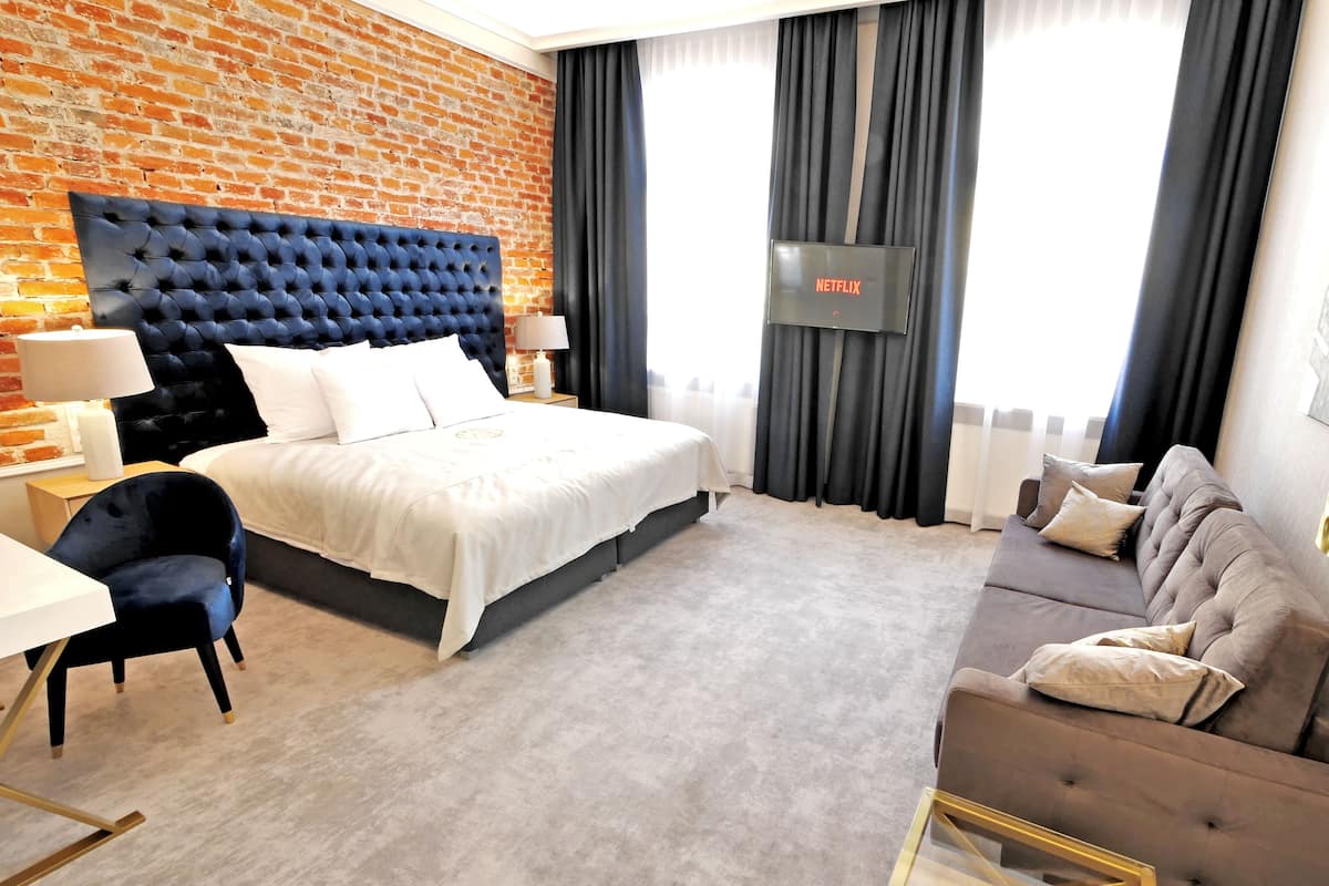 Una habitación de hotel con pared de ladrillo, cama con cabecera capitonada, sofá gris, escritorio y televisor montado en la pared.
