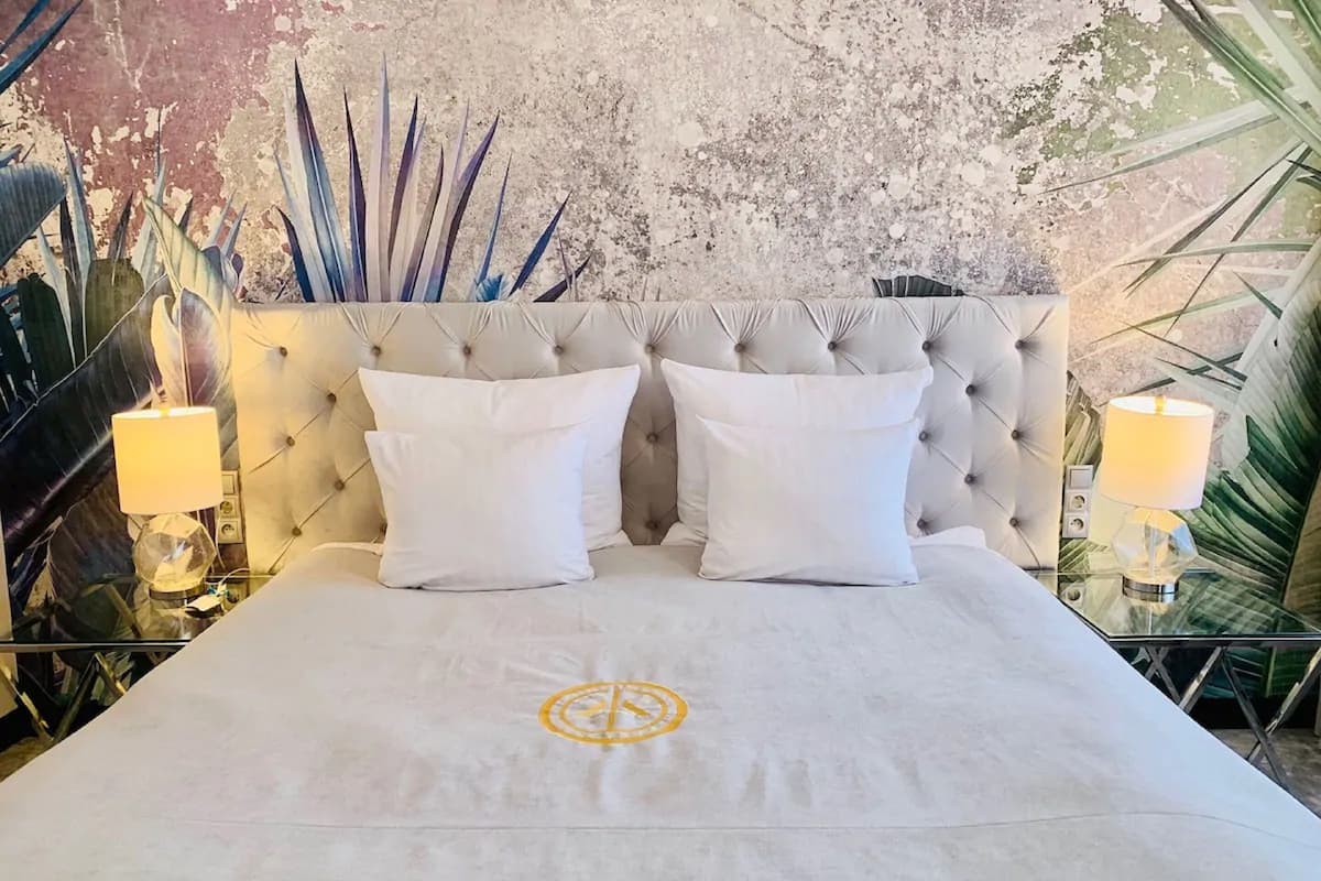 Una cama con cabecera capitonada, dos almohadas blancas y una colcha con un emblema circular.