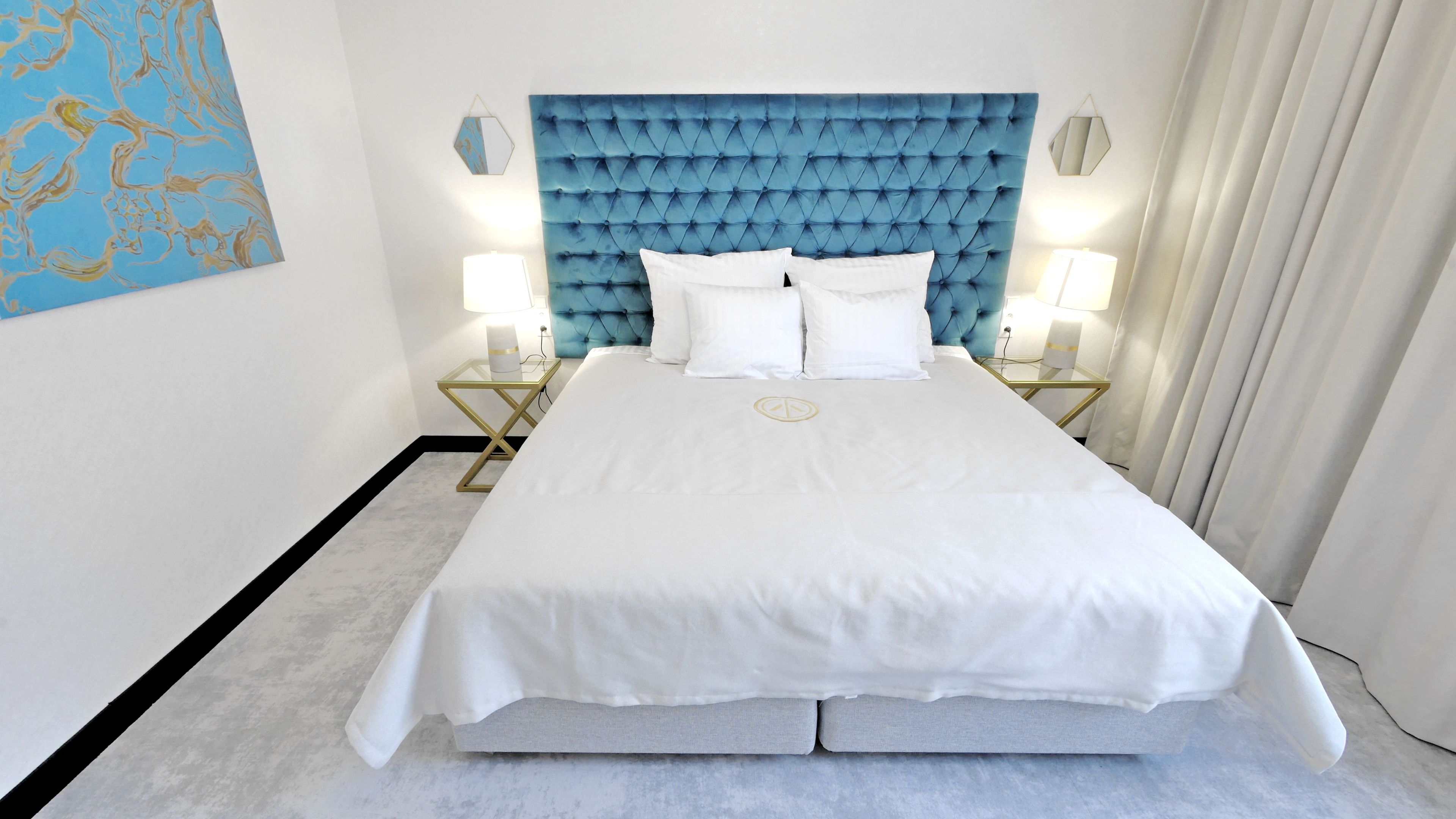 A hotel room with a large bed, two bedside tables with lamps, a blue tufted headboard, and a white bedspread.