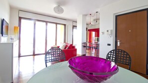 Un salon moderne avec un canapé rouge, une table basse en verre surmontée d’un bol violet, et un coin repas avec deux chaises.