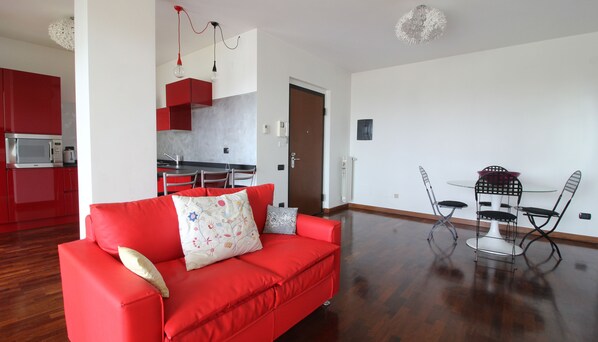 Een moderne woonkamer met een rode bank, een eethoek met een glazen tafel en metalen stoelen, en een keuken met rode kasten en een magnetron.