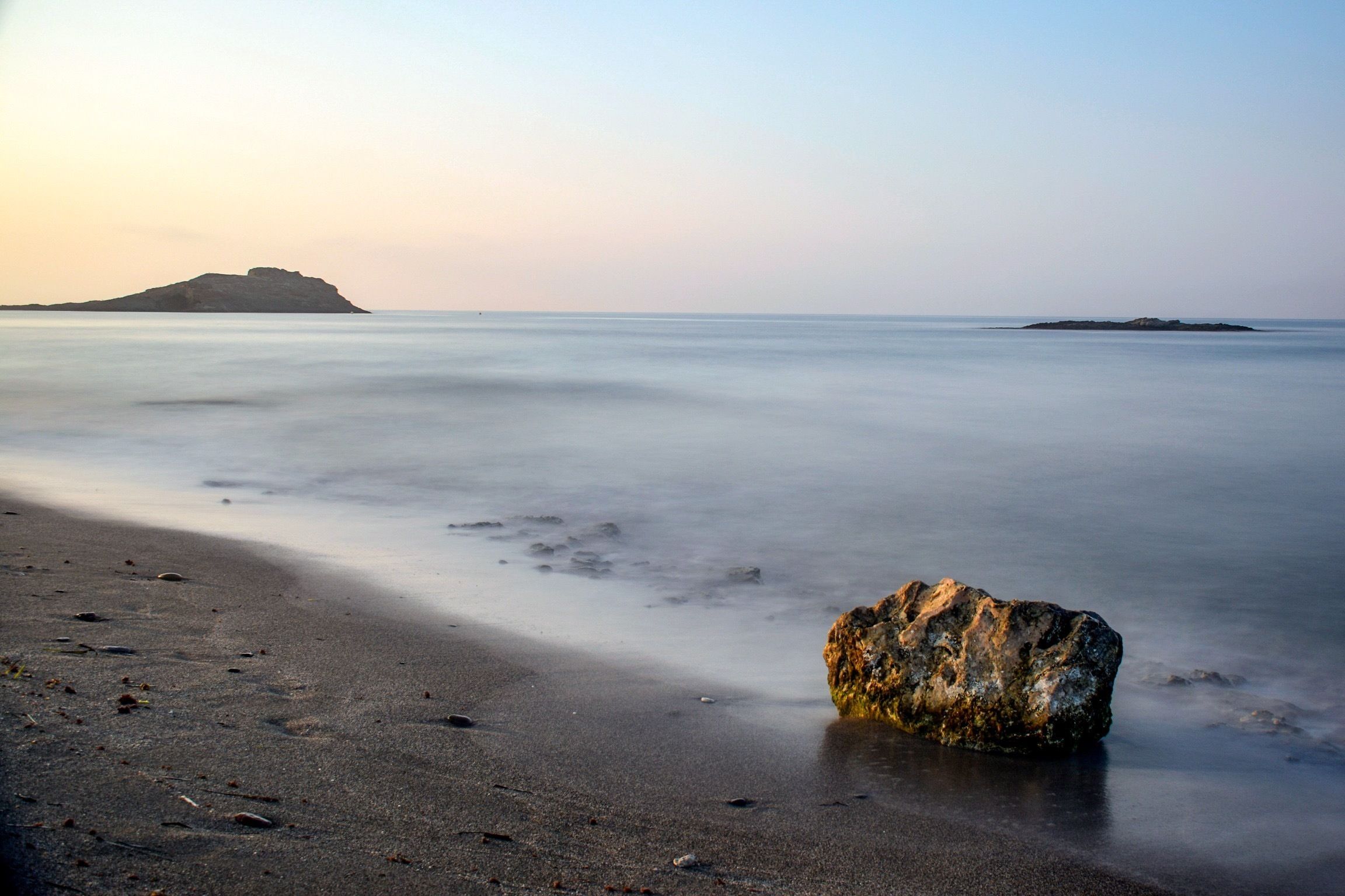 A beach with a single rock in the foreground and calm water extending to distant landforms.