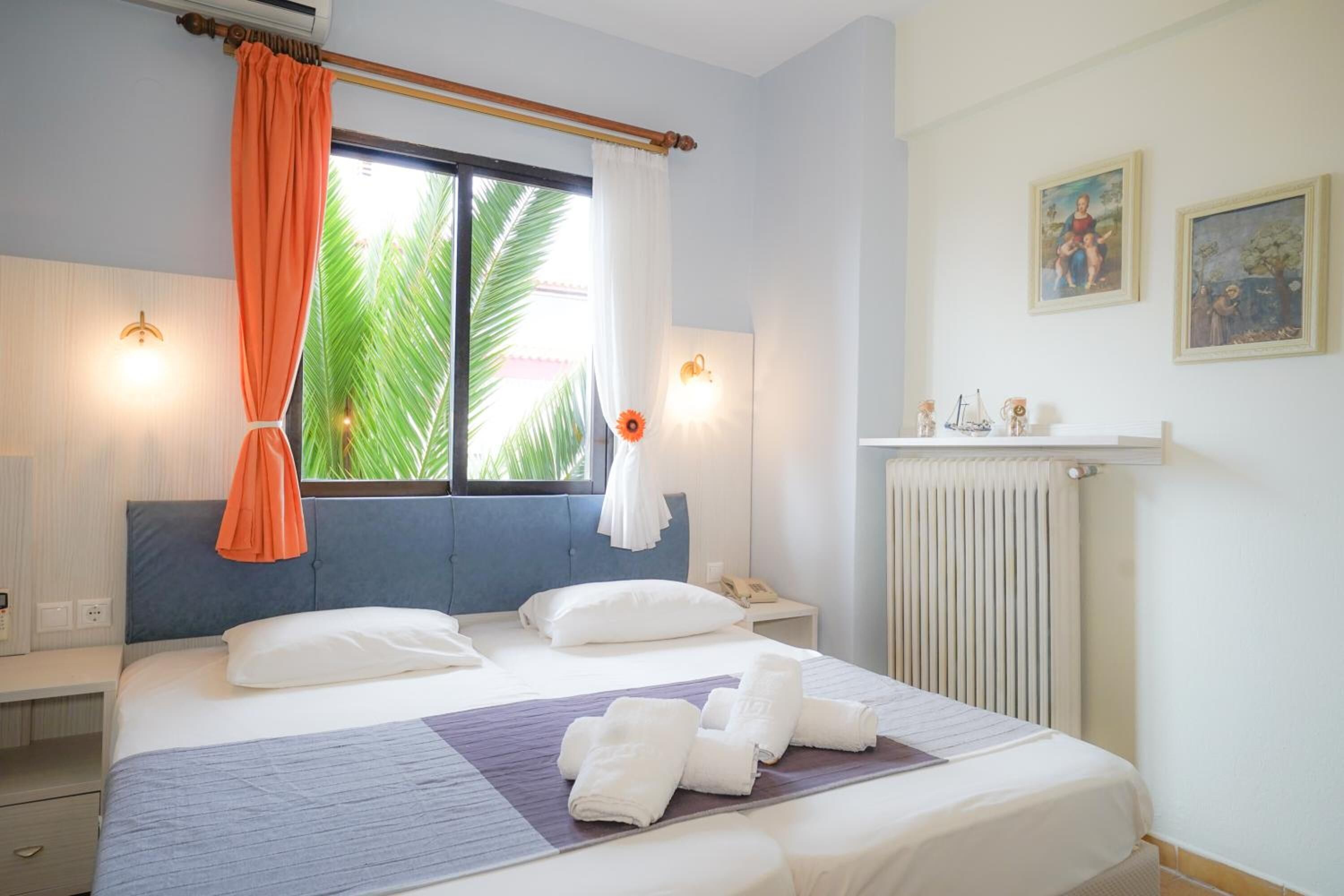 A hotel room with a bed, a window with curtains, a bedside table with a lamp, a nightstand, and a radiator.