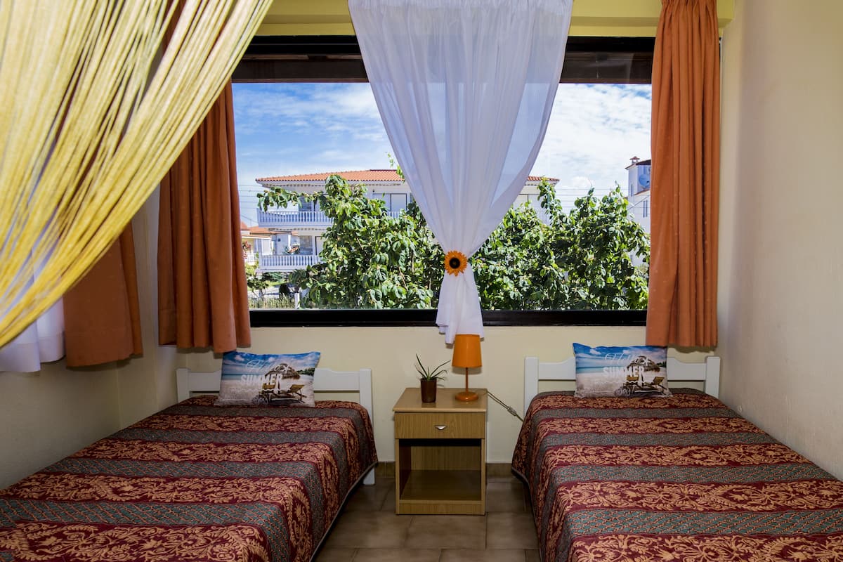 Un dormitorio con dos camas, una mesita de noche, una ventana con cortinas y vistas a edificios y árboles.