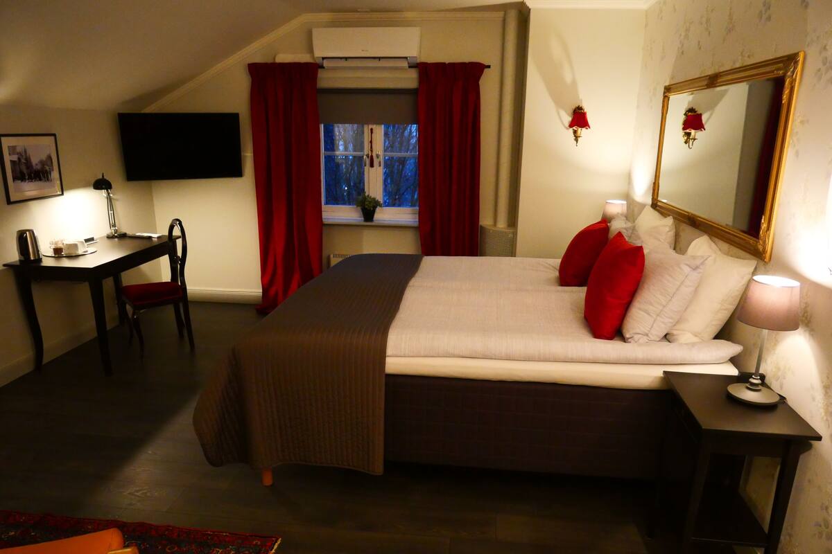 A bedroom with a bed, a desk, a chair, a lamp, a mirror, and a window with curtains.