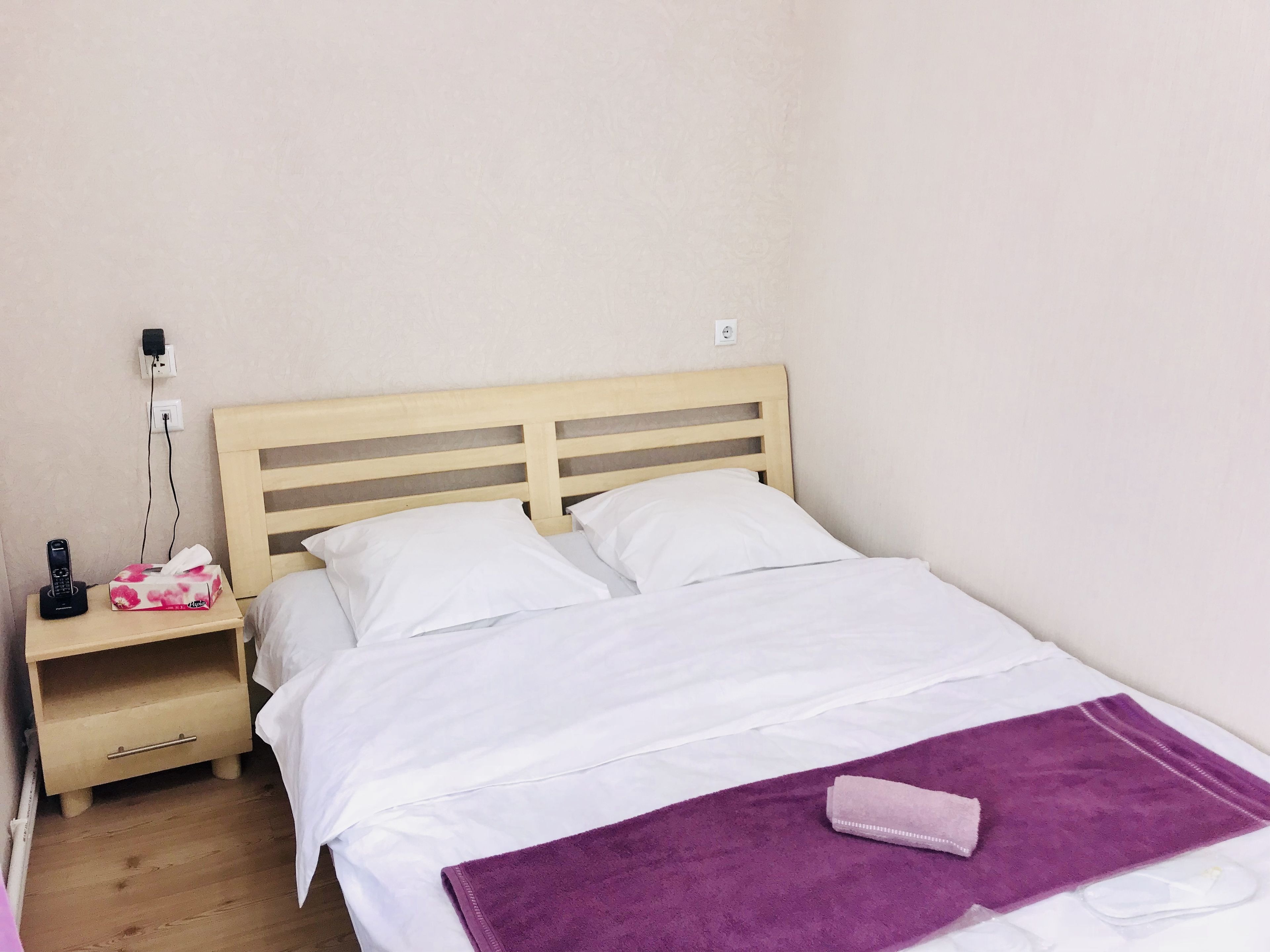 Un letto singolo con testiera in legno, biancheria da letto bianca, un asciugamano viola e un comodino con una scatola di fazzoletti e un caricabatterie per telefono.