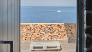 A view from a balcony with a stone wall, a white bench, and a sailboat on the water. - Cape Suites (Kithnos)