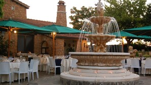 Un espace de restauration en plein air avec des tables et des chaises en pierre, une fontaine en pierre et des parasols verts.