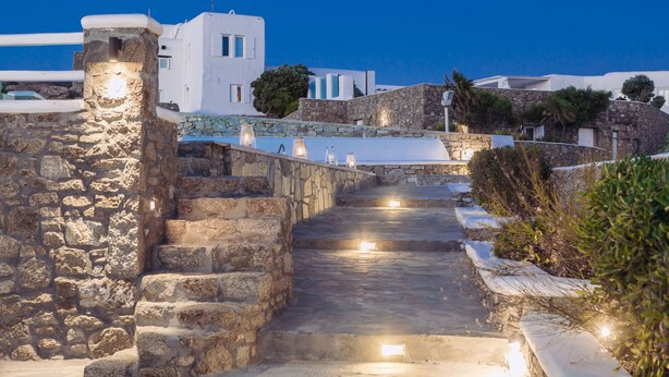 A stone staircase leading to a white building with a flag, surrounded by greenery and stone walls. - Villa Mando (Mykonos)