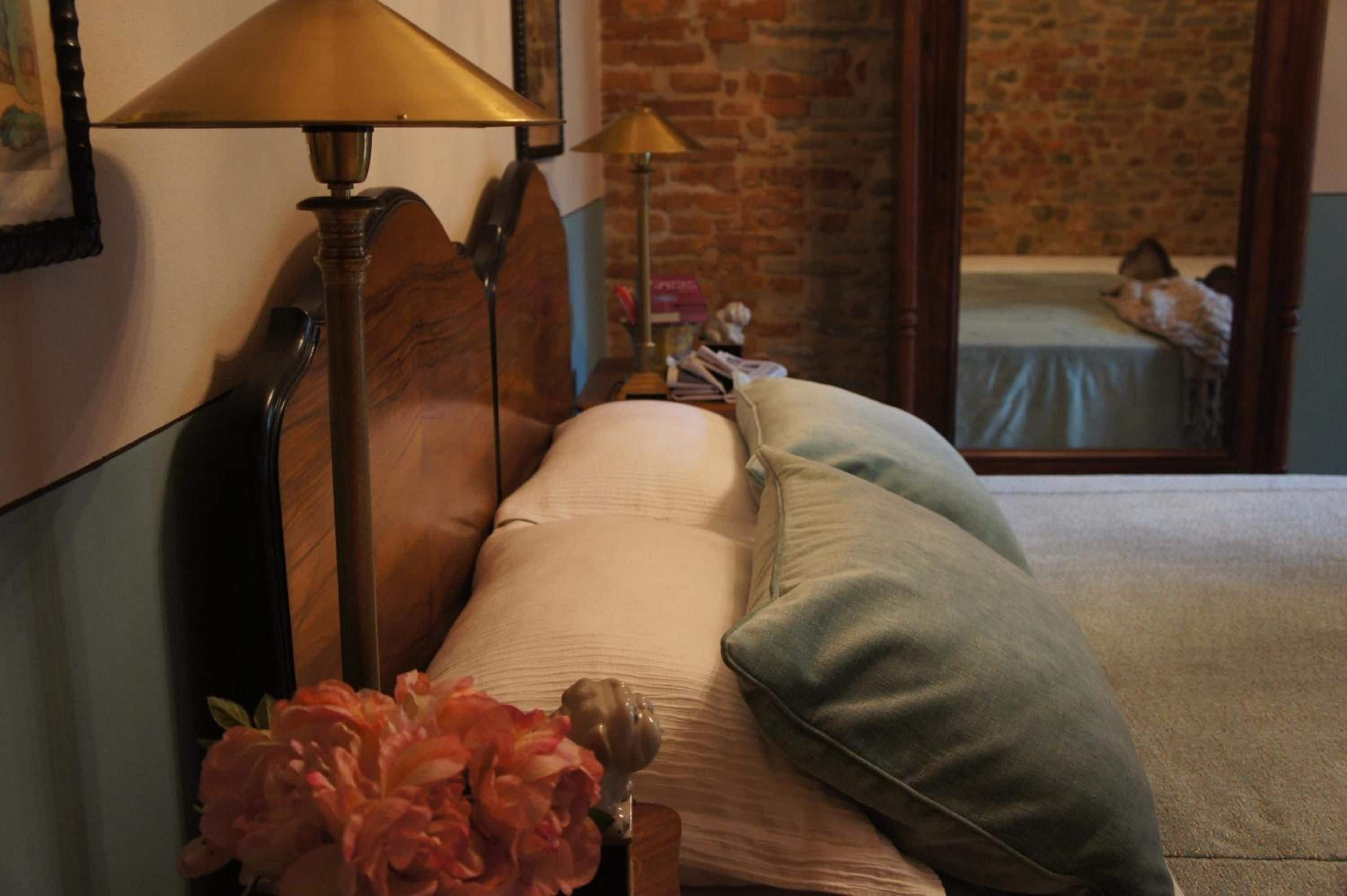 A bed with pillows, a lamp, and a vase of flowers in a room with a brick wall and a mirror reflecting a bed.