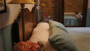A bed with pillows, a lamp, and a vase of flowers in a room with a brick wall and a mirror reflecting a bed.