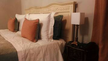 Una cama bien hecha con una colcha blanca y varias almohadas, una lámpara de noche y una mesita de noche de madera oscura con un teléfono.
