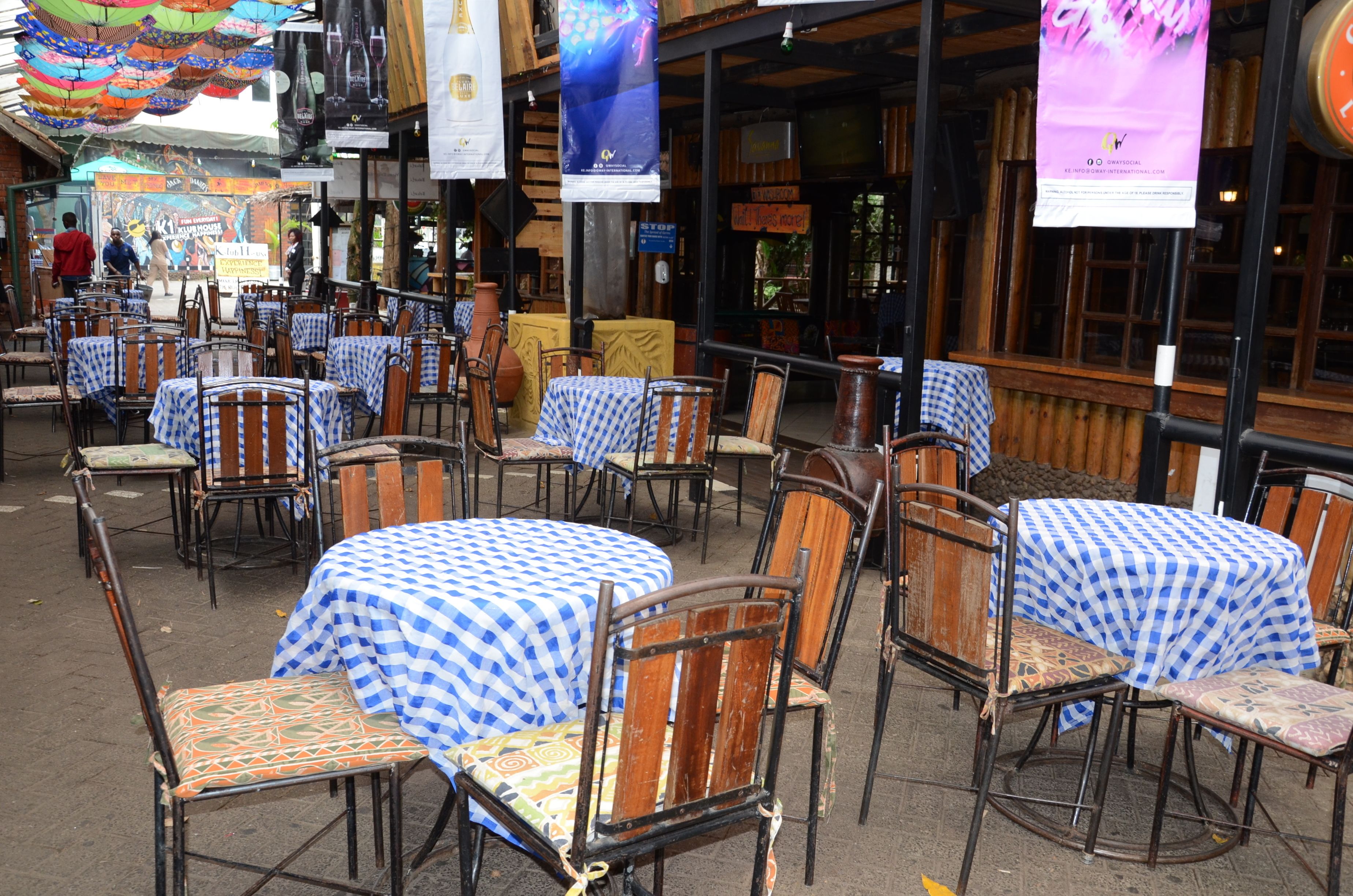 An outdoor cafe with tables covered in blue and white checkered tablecloths, wooden chairs, and hanging decorations.