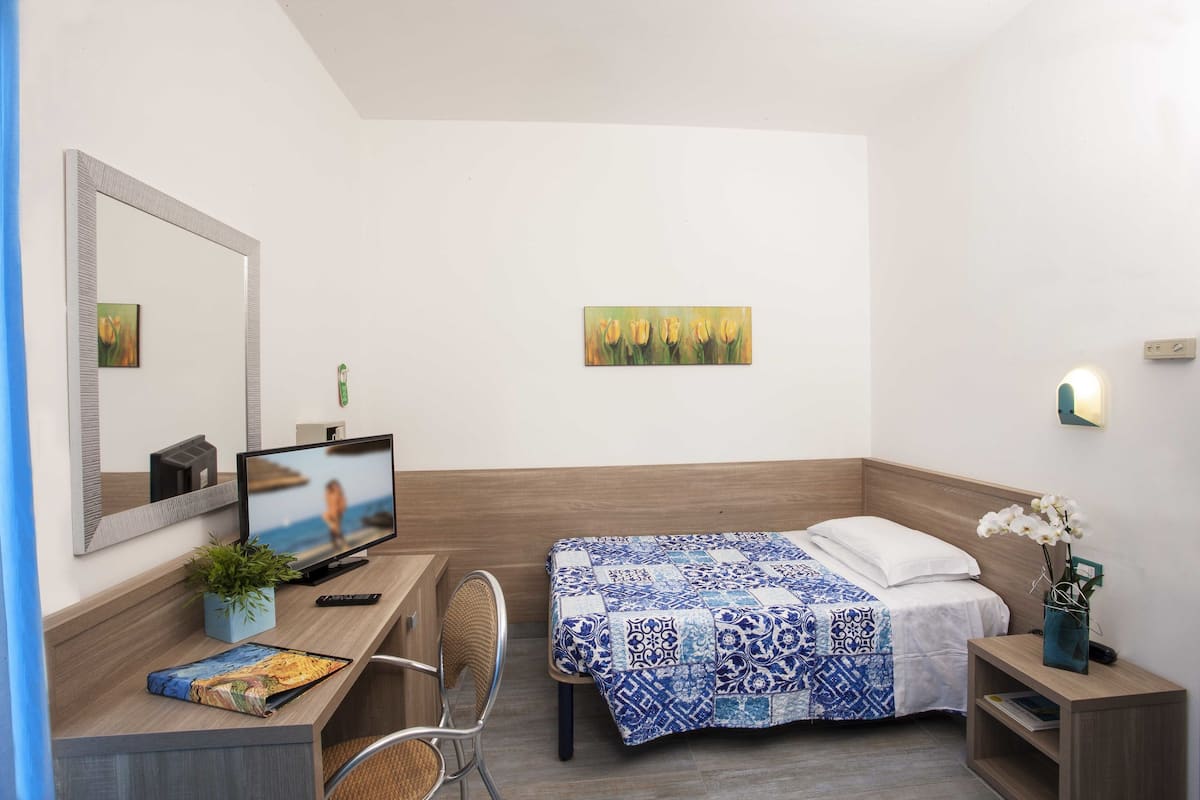 A hotel room with a bed, a desk with a computer, a chair, a TV, and a painting on the wall.