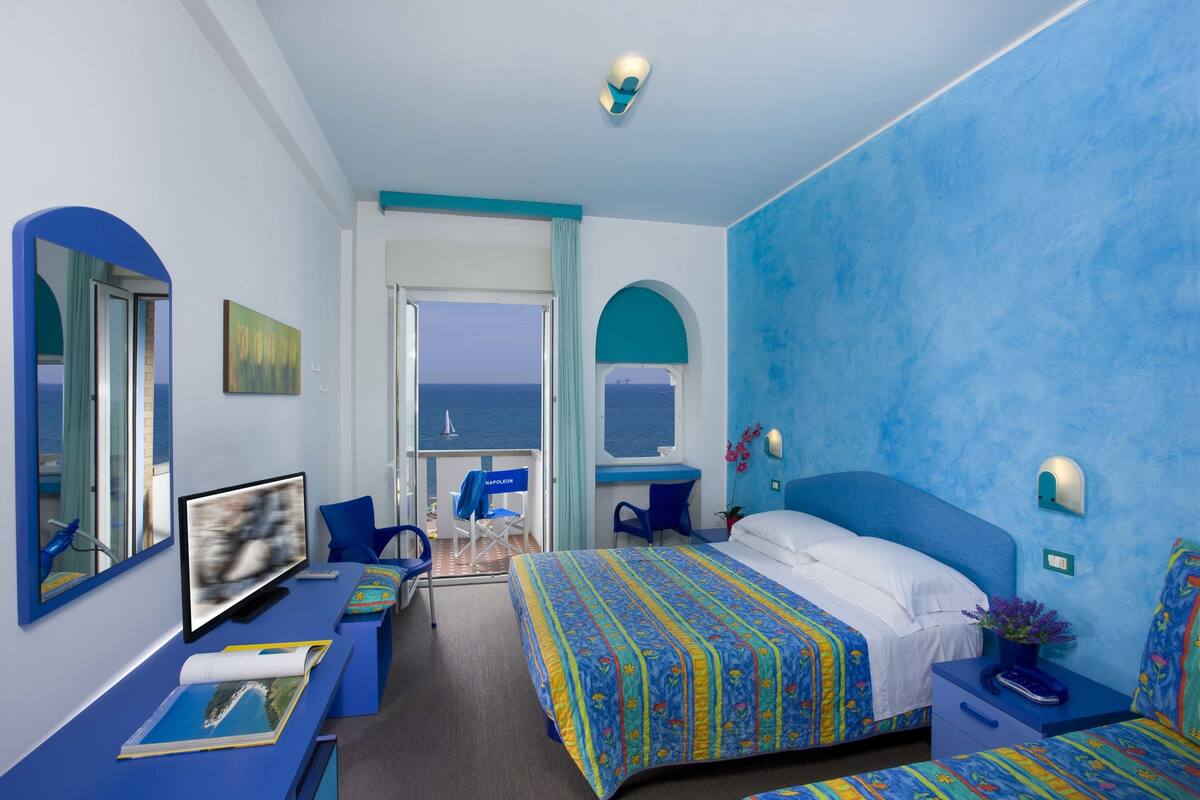A hotel room with a large bed, a desk with a computer, a chair, and a view of the sea.
