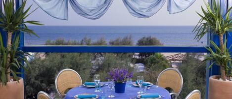 A dining table set for six with a blue tablecloth, surrounded by wicker chairs, overlooking a scenic ocean view.