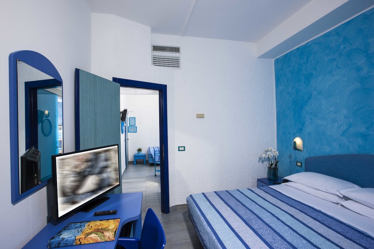 A hotel room with a bed, a desk with a computer, a TV, and a bathroom visible through an open door.
