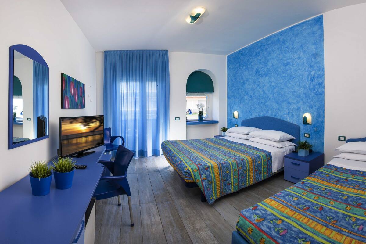 A hotel room with a desk, two beds, a large window with blue curtains, and a blue accent wall.