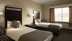 A hotel room with two beds, a nightstand, a lamp, a chair, and a window with curtains. - Wessex Hotel (Street)