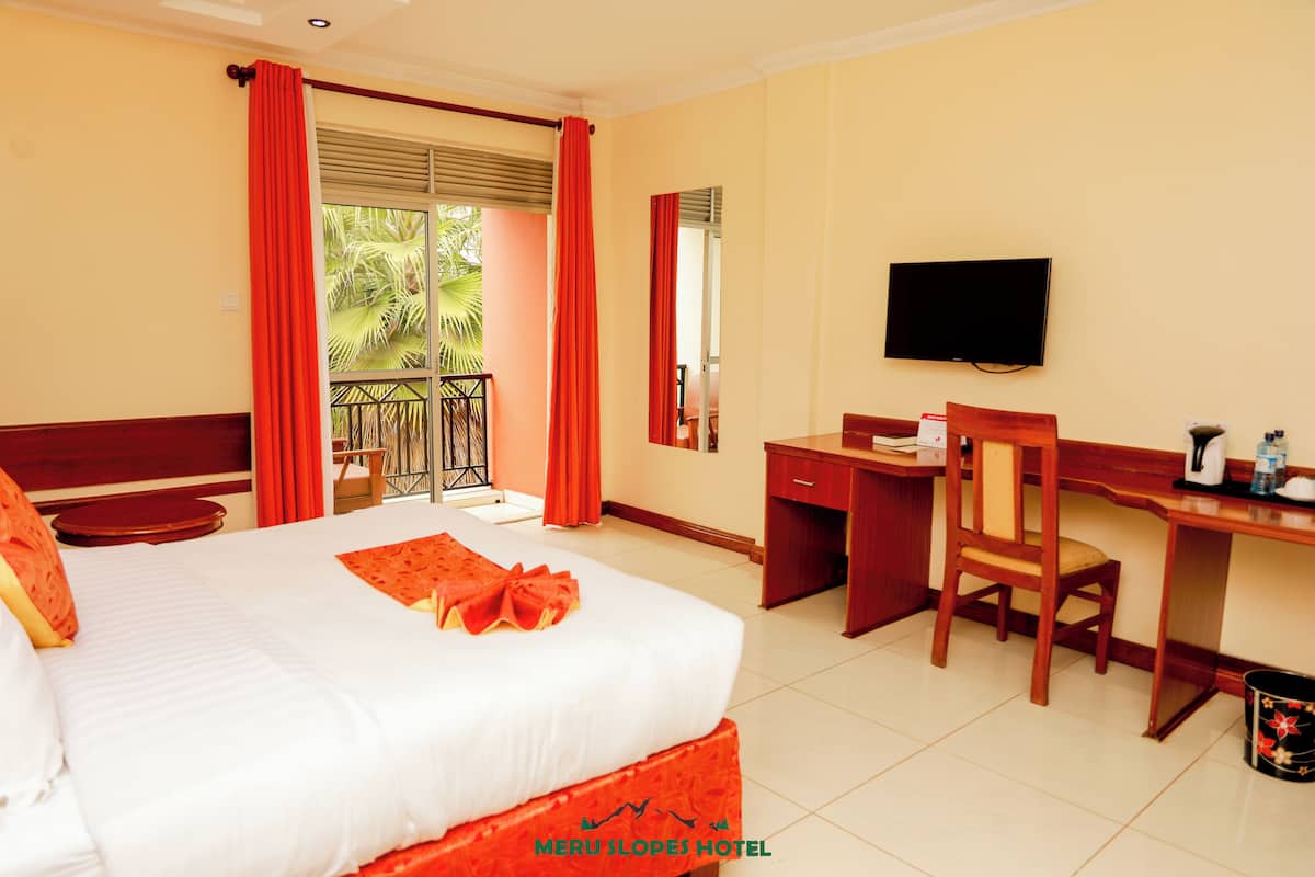 Habitación de hotel con cama, escritorio, silla, televisión y balcón con cortinas rojas.