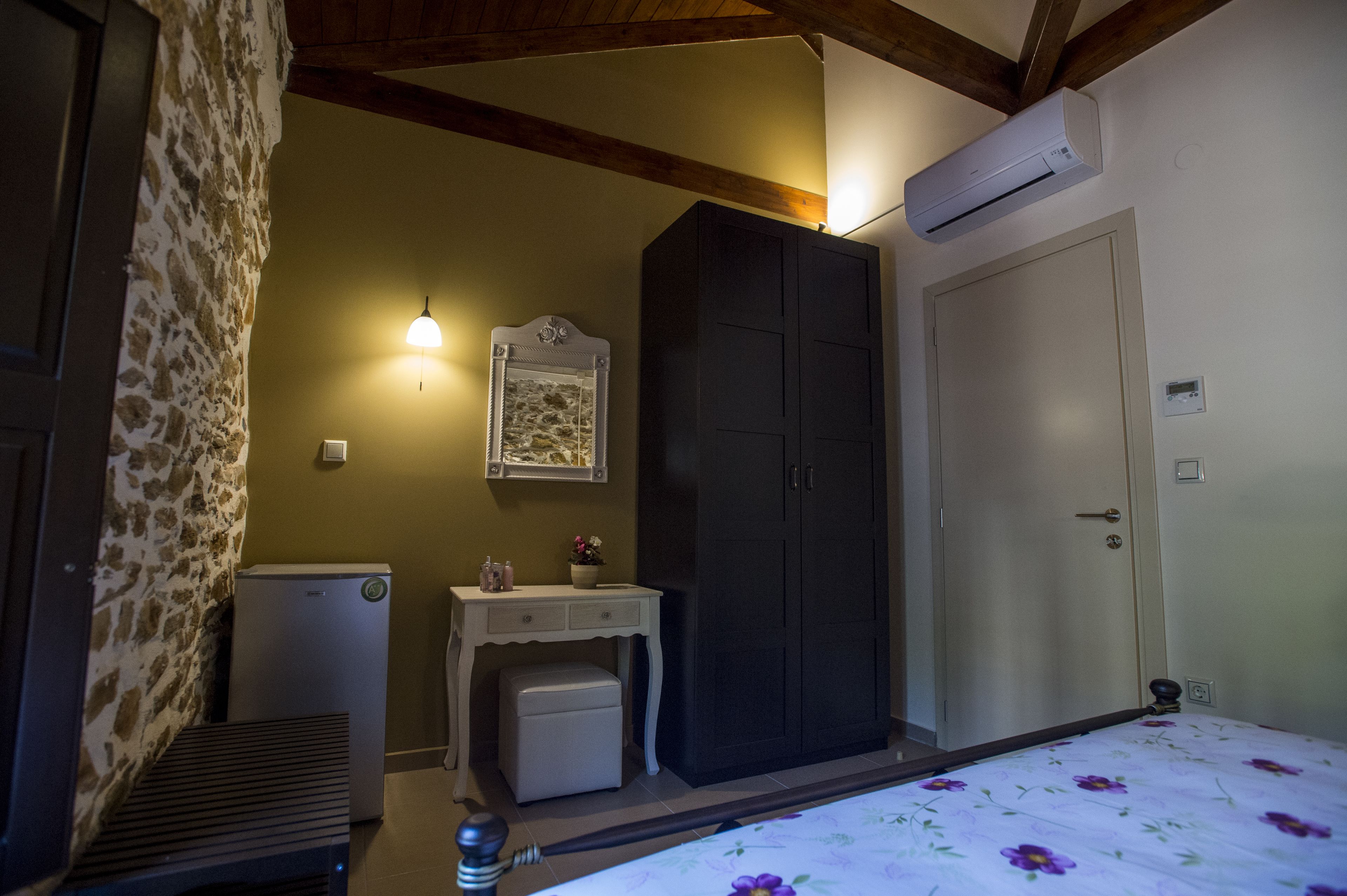 A bedroom with a bed, a wardrobe, a small desk with a mirror, and an air conditioning unit.