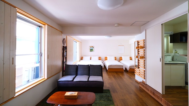 A compact hotel room with a sofa, a small table, a bed with two pillows, a bedside table, a window, and a kitchenette. - Rooms (Tokyo)