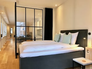A modern bedroom with a large bed, a bedside table, and a view through a glass partition to a dining area.