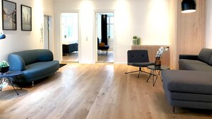 Una sala de estar moderna con un sofá, sillones y una mesa de centro. Hay cuadros enmarcados en la pared, una planta en maceta y un radiador.