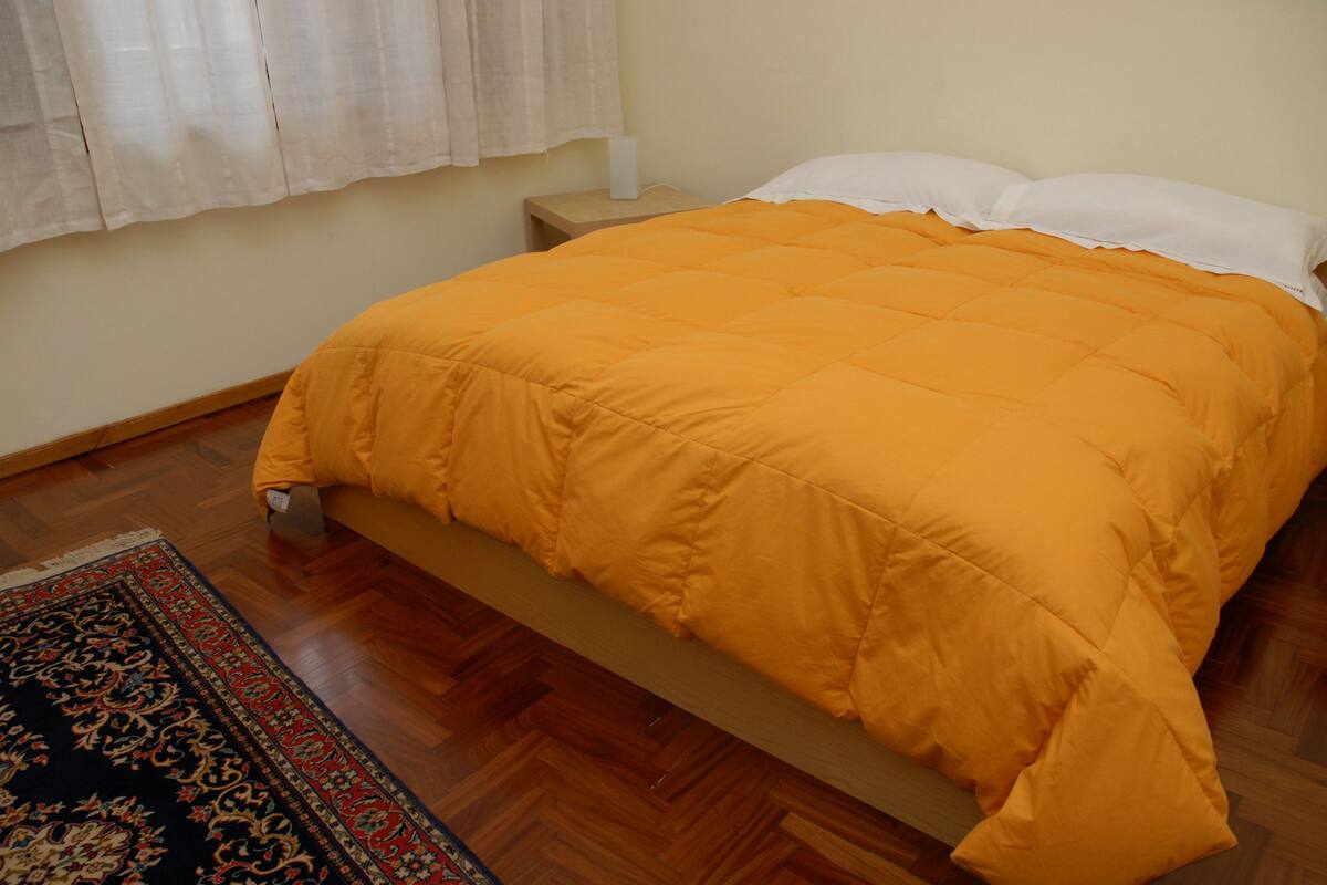A bed with an orange blanket, a wooden floor, and a patterned rug.