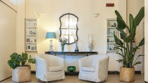 A room with two white armchairs, a table with a lamp, a mirror, and a large green plant. - Hotel Massimo (Cecina)