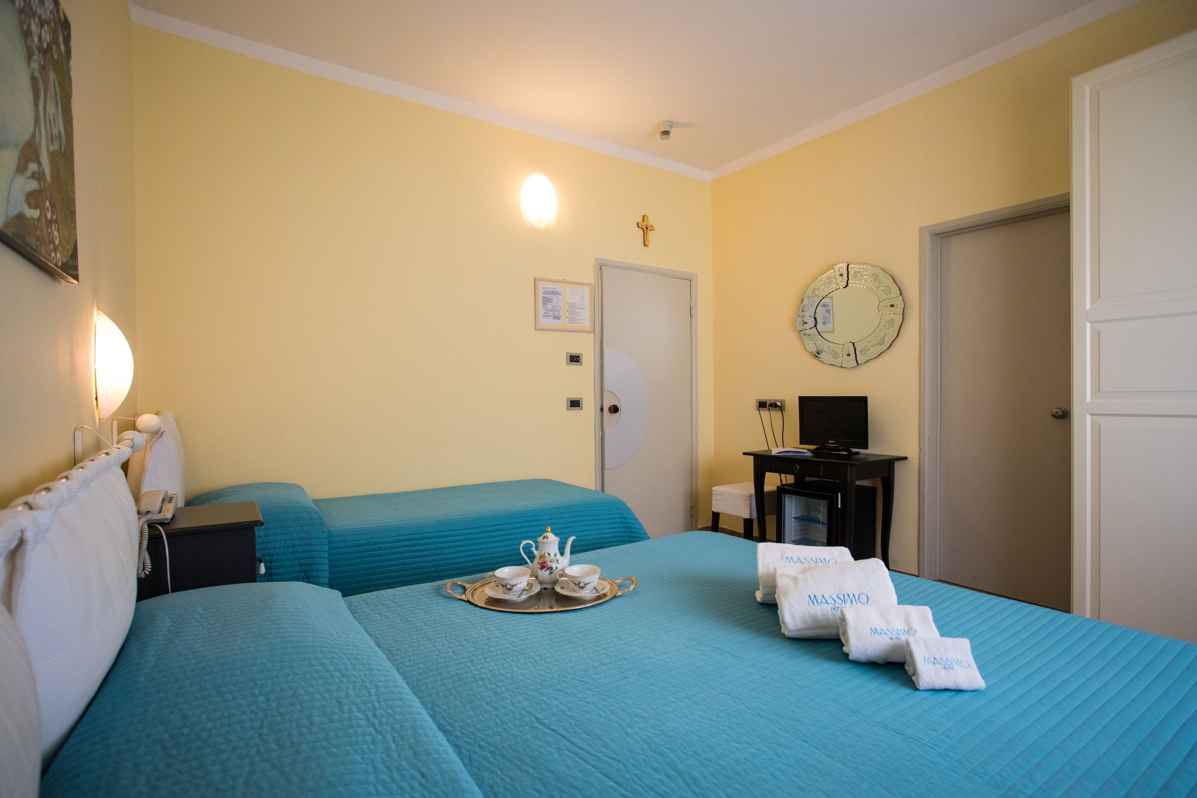 A hotel room with two beds, a desk with a computer, a clock, and a door.