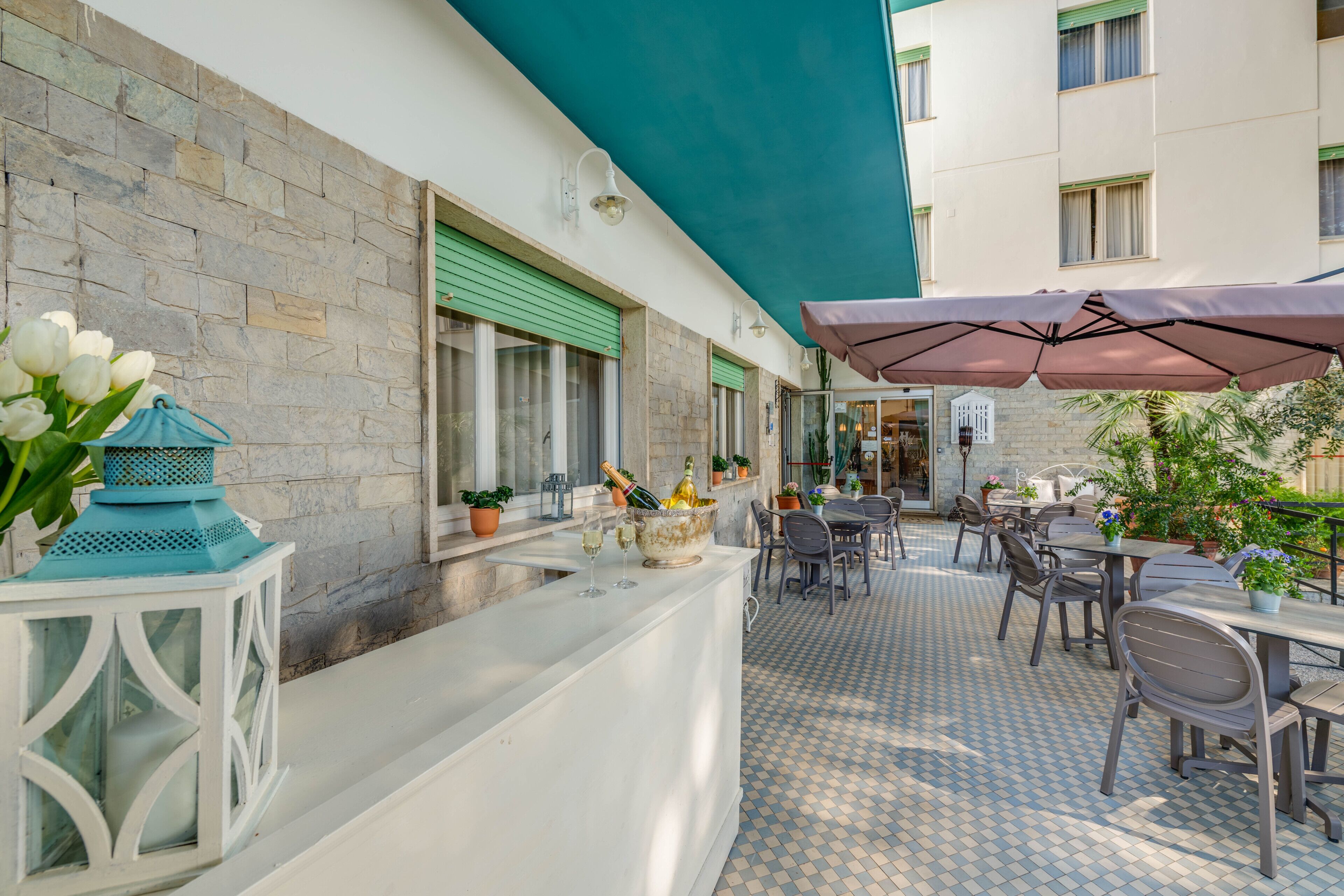 An outdoor bar area with a stone wall, green shutters, and a tiled floor. There are tables and chairs under a large umbrella, and a white lantern with flowers is visible.
