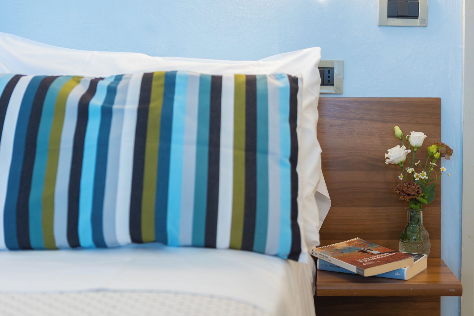 A neatly made bed with a striped pillow, a small bedside table with a book and a vase of flowers, and a wall socket.