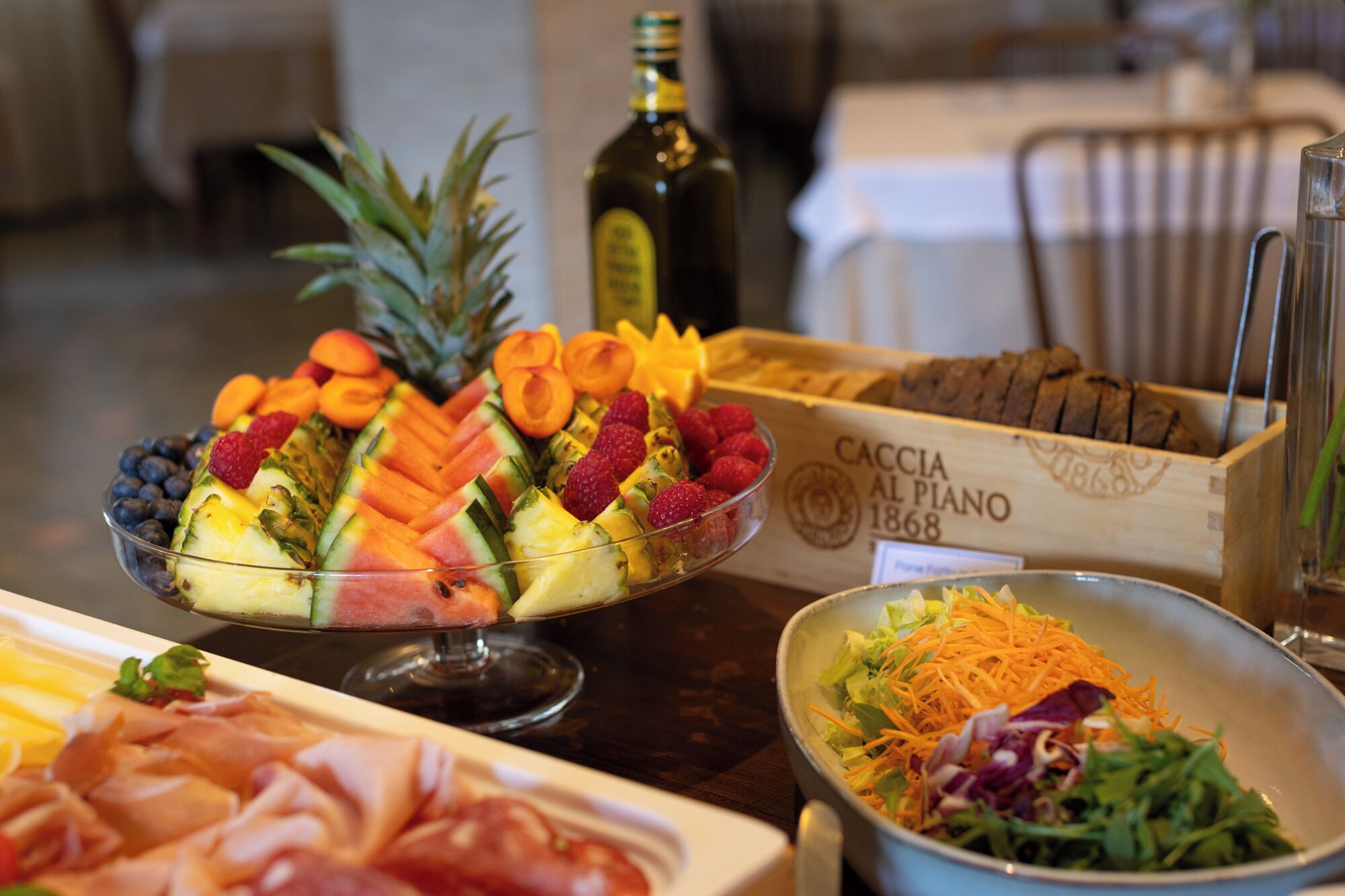 A table with a fruit salad, a box labeled 'CACCIA AL PIANO 1868', and a salad bowl.