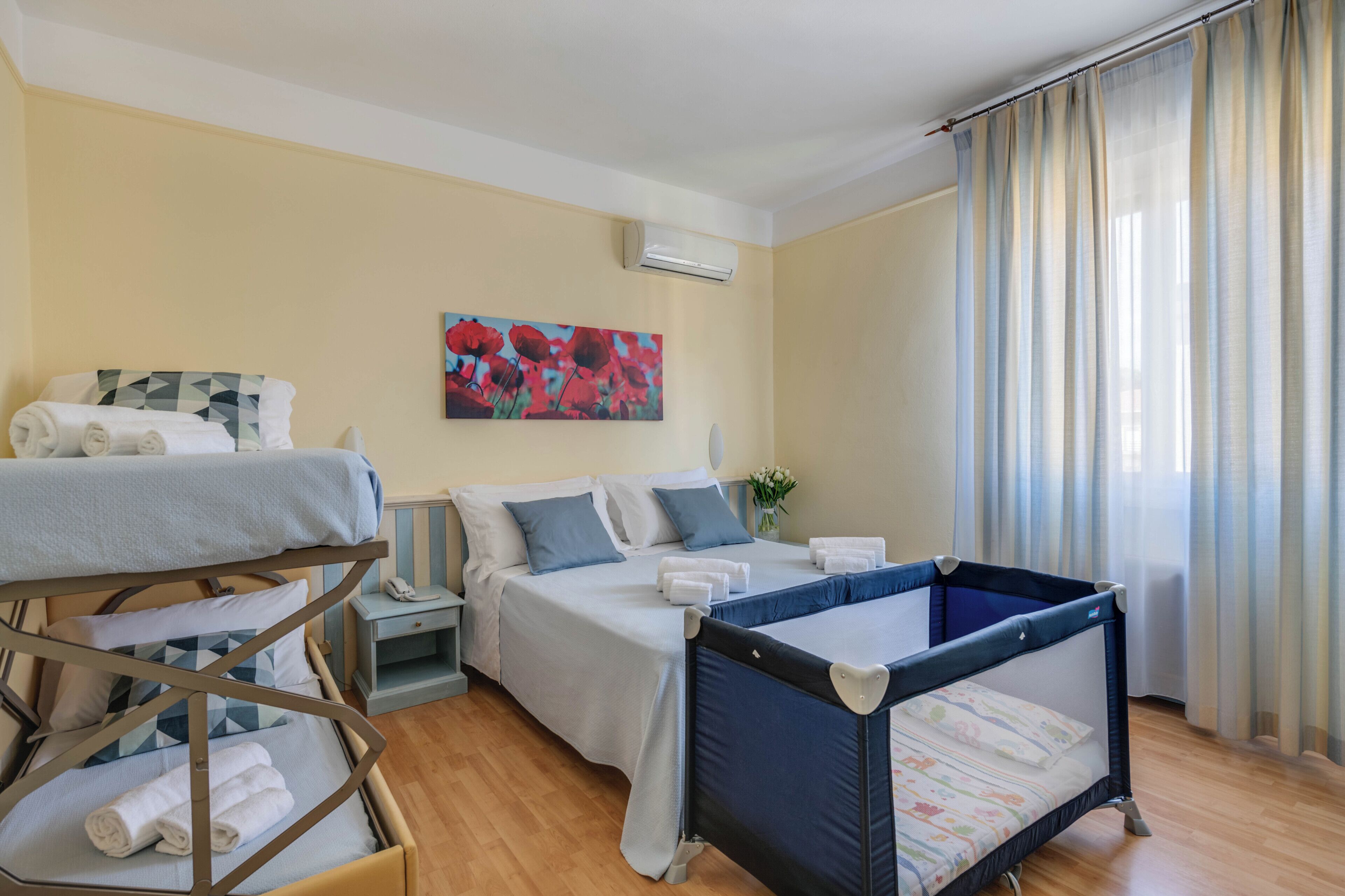 A hotel room with a bunk bed, a single bed, a crib, a painting, an air conditioning unit, and a window with curtains.
