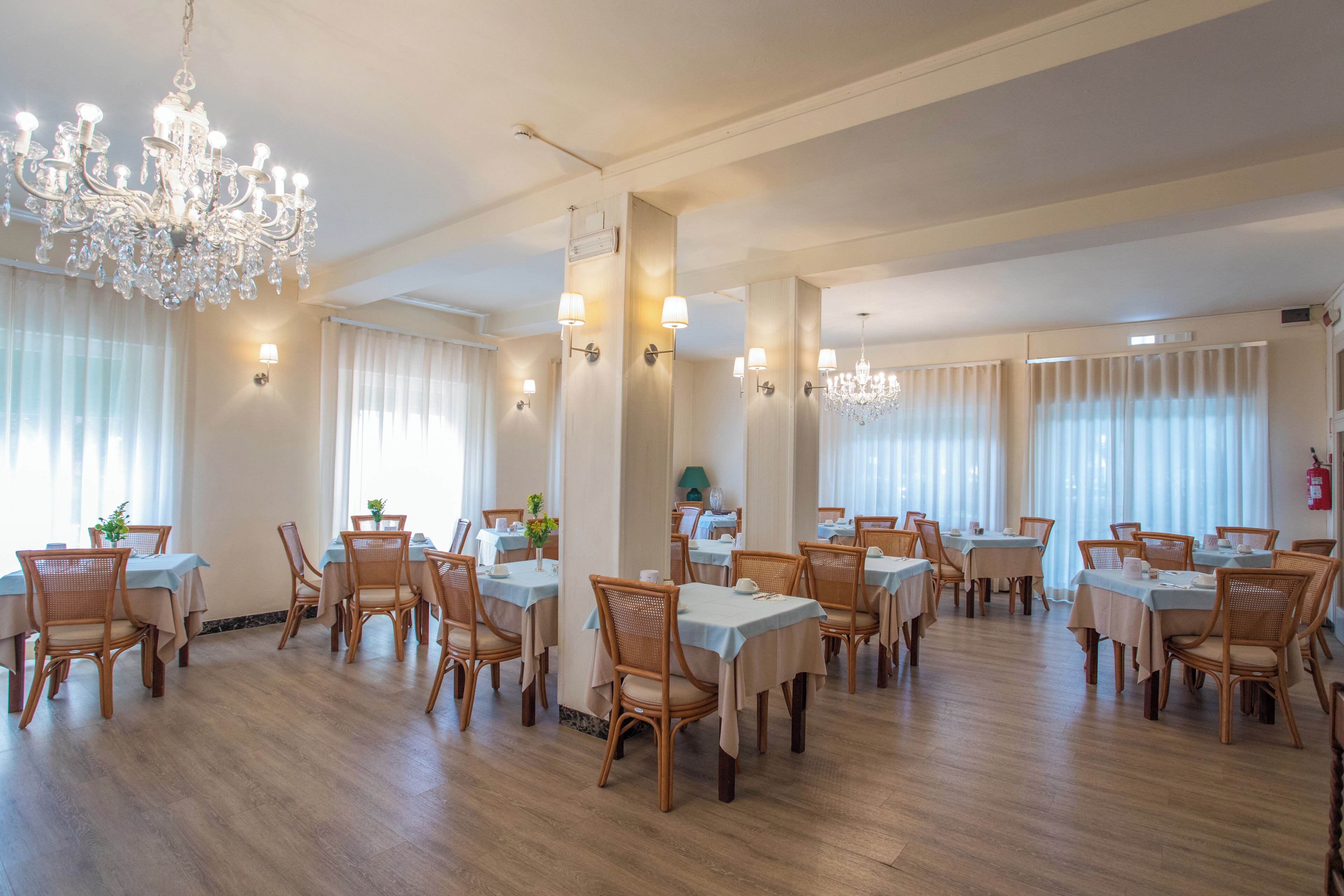 A spacious dining area with multiple tables and chairs, elegant chandeliers, and large windows with sheer curtains.